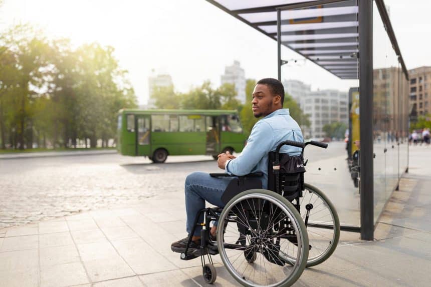 Young disabled black man in wheelchair waiting for public transport on bus stop, having difficulty