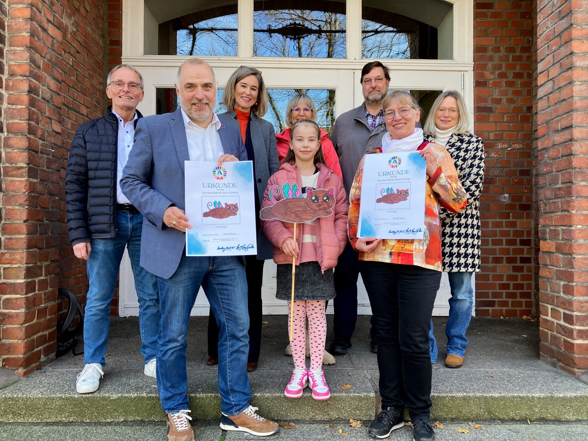 Meinolf Padberg, Projektleiter „DemokraTIER“ an der Roten Schule, Bürgermeister Ralf Paul Bittner, Esther von Kuczkowski, Leiterin des Dezernates Bildung|Kultur|Sport der Stadt Arnsberg, Julia Teasdale und Michael Froebus vom Bürger:innenrat Arnsberg, Schulleiterin Thekla Bock-Weitershagen und Kirsten Heckmann, Leiterin des Bürgermeisterreferates, gratulieren Nicole Block aus der Klasse 4a der „Roten Schule“ in Neheim zum Sieger-Entwurf für das „DemokraTIER“ der Stadt Arnsberg. (Foto: Stadt Arnsberg)